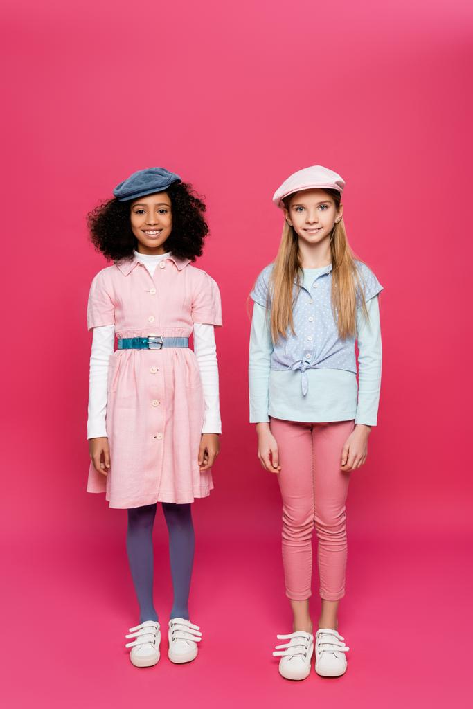 volledige weergave van twee gelukkig interraciale meisjes in trendy kleding poseren op roze - Foto, afbeelding