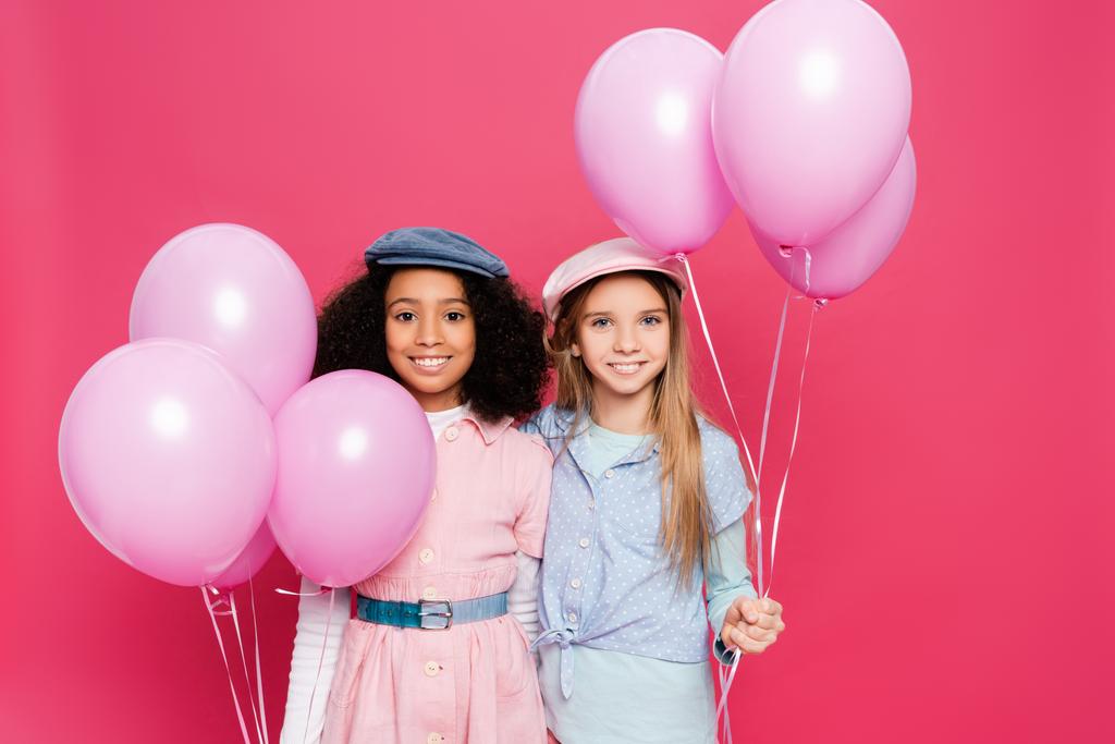 δύο χαρούμενα πολυπολιτισμικά κορίτσια με μοντέρνα ανοιξιάτικα ρούχα ποζάρουν με γιορτινά μπαλόνια απομονωμένα σε ροζ  - Φωτογραφία, εικόνα