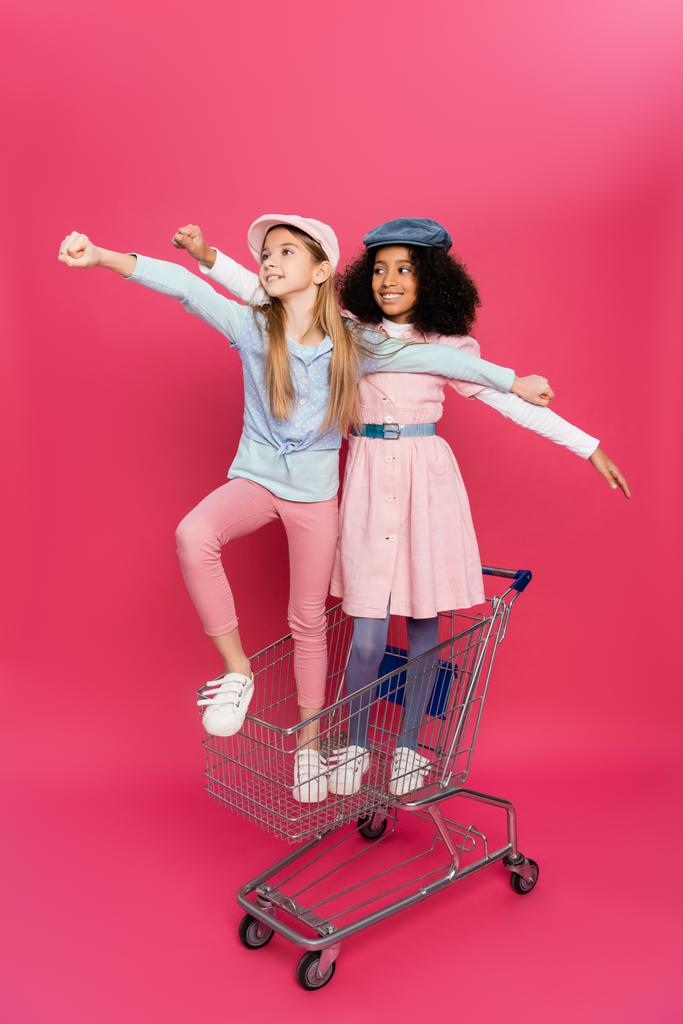 alla moda, felici ragazze interrazziali in posa nel carrello della spesa su rosa - Foto, immagini