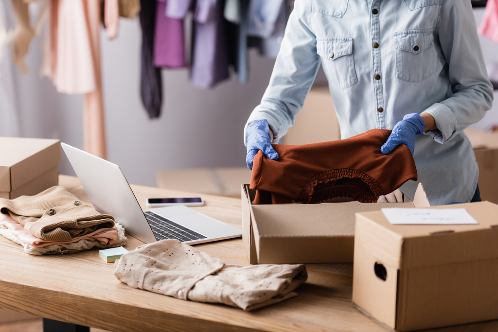 Ausgeschnittene Ansicht eines Verkäufers in Latexhandschuhen, der Kleidung in Schachteln in einer Modeboutique verpackt - Foto, Bild