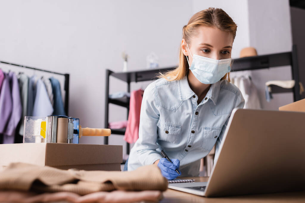 fiatal bemutatóterem tulajdonosa orvosi maszk, közel dolgozik laptop elmosódott előtérben - Fotó, kép