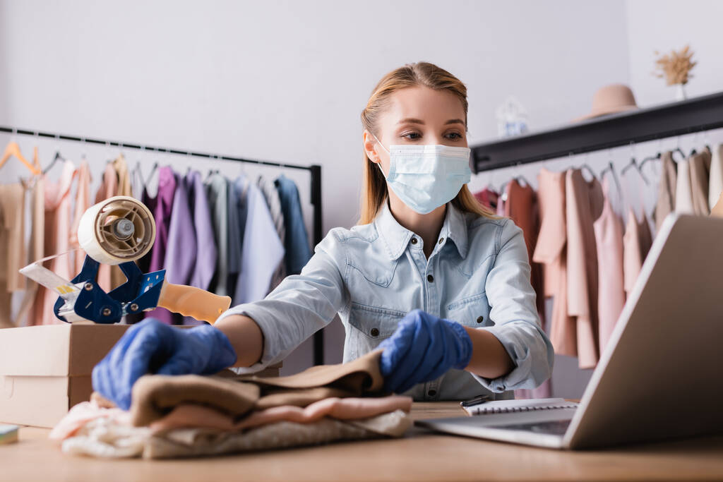 mladý prodejce v lékařské masce, práce s oděvy v blízkosti notebooku v showroomu na rozmazaném popředí - Fotografie, Obrázek