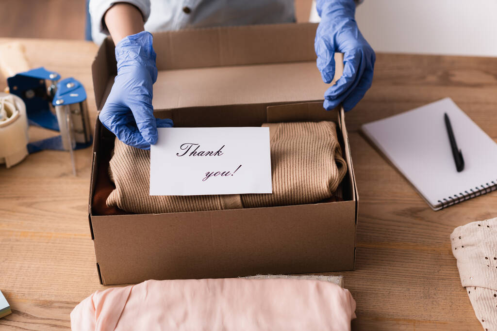 Ausgeschnittene Ansicht des Showroom-Besitzers in Latexhandschuhen Verpackungskarte mit Dankeschön-Aufschrift in Schachtel, verschwommener Hintergrund - Foto, Bild