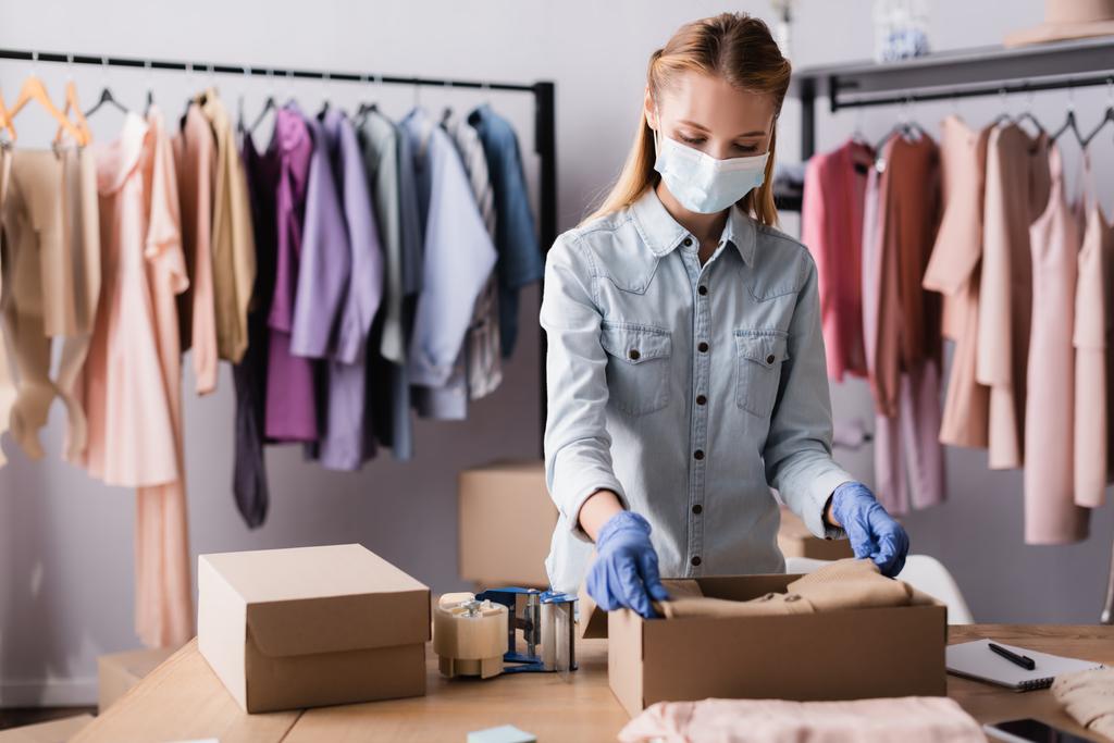 молодая бизнесвумен в медицинской маске, упаковывает одежду в коробки в выставочном зале, размытый передний план - Фото, изображение