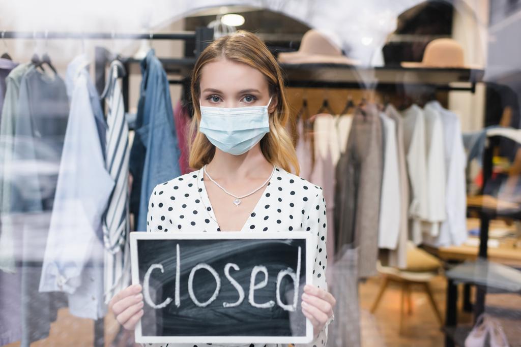showroom owner in medical mask, looking at camera and holding board with closed lettering  - Photo, Image