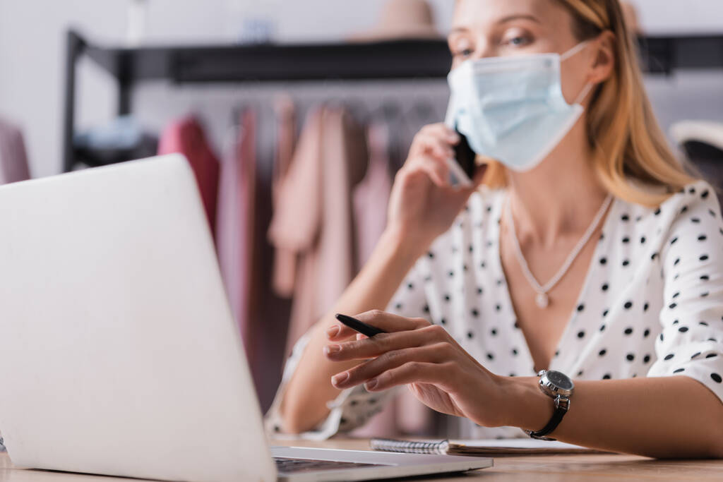 бизнесвумен в медицинской маске принимает заказ в выставочном зале, работая рядом с ноутбуком на размытом фоне - Фото, изображение