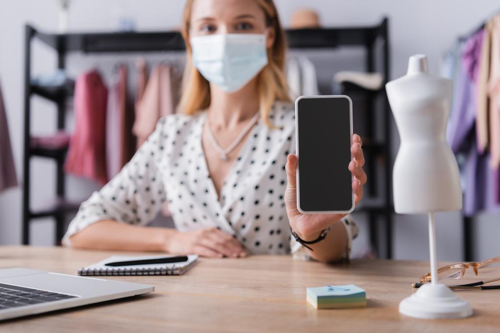 Geschäftsfrau in medizinischer Maske, zeigt Handy mit leerem Bildschirm, während sie im Showroom auf verschwommenem Hintergrund arbeitet - Foto, Bild