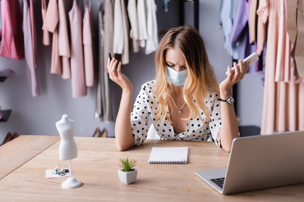 frowning businesswoman in medical mask, holding smartphone and pen near blank notebook in showroom - Photo, Image