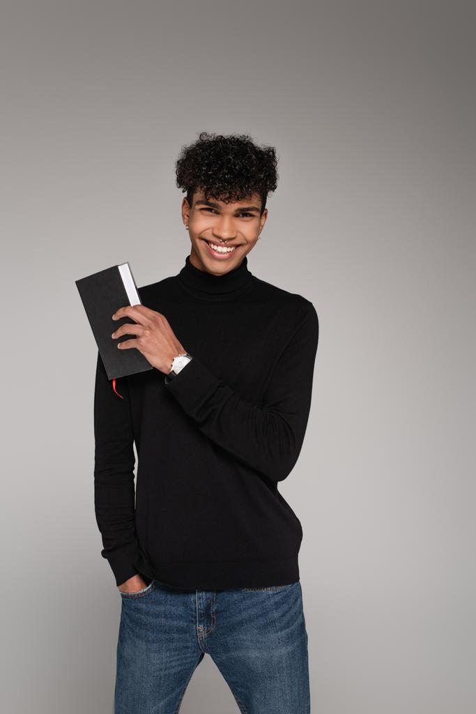 cheerful african american man in turtleneck sweater holding notepad isolated on grey - Photo, Image