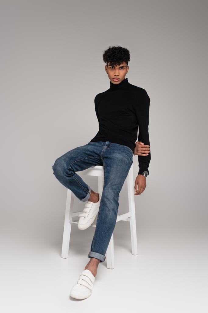 full length of young african american man sitting on chair while posing on grey - Photo, Image