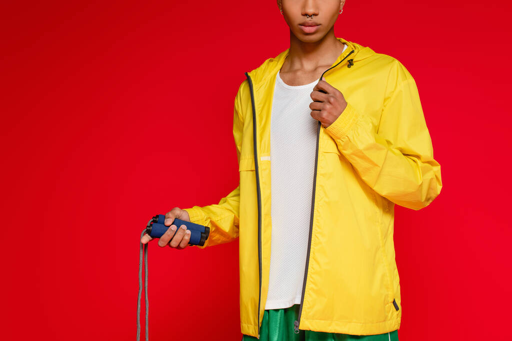 recortado vista de deportivo africano americano hombre en amarillo chaqueta de lluvia celebración saltar cuerda aislado en rojo - Foto, Imagen