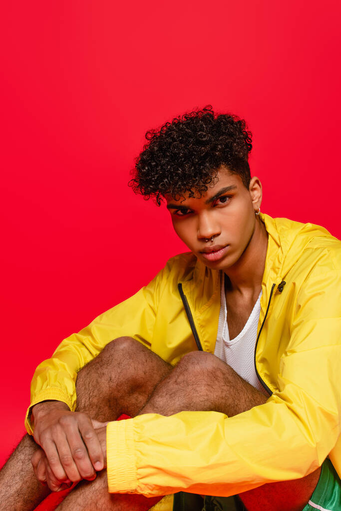 pierced african american man in yellow jacket sitting while posing isolated on red - Photo, Image