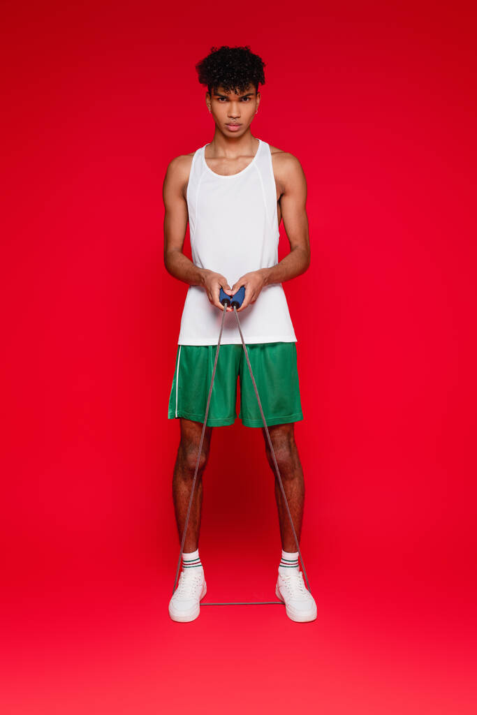 full length of sportive african american man in shorts and tank top exercising with skipping rope on red - Photo, Image