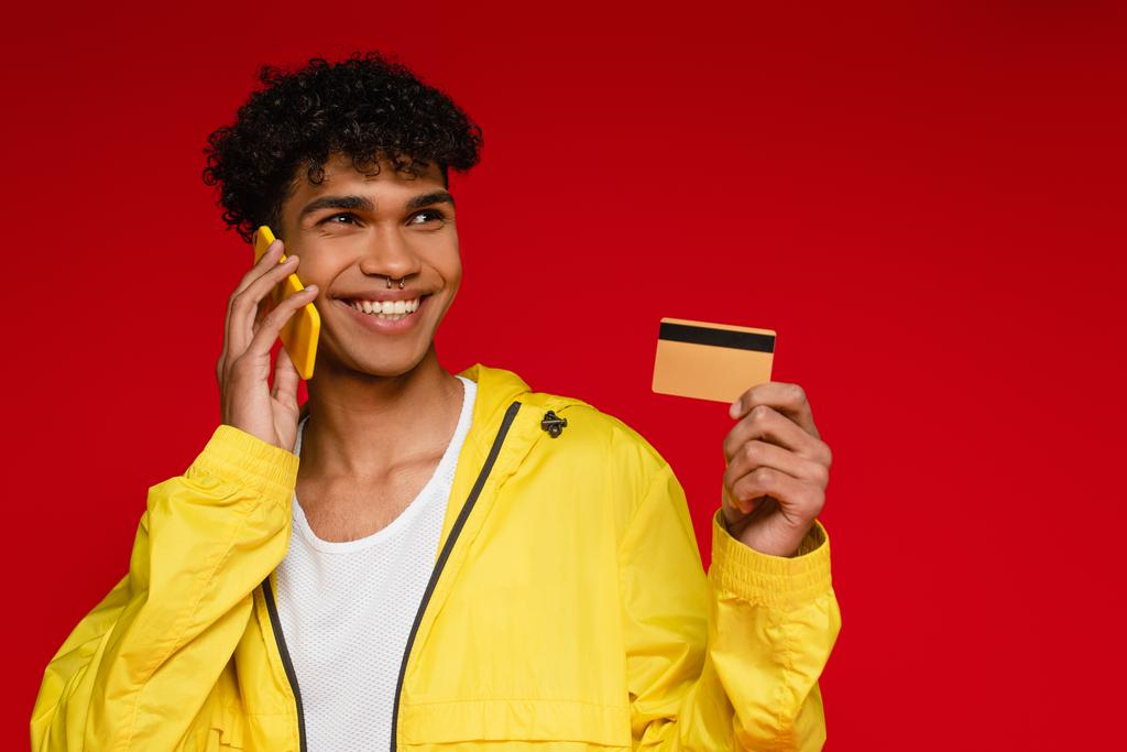 gelukkig Afrikaans amerikaanse man in jas praten op smartphone en houden credit card geïsoleerd op rood - Foto, afbeelding