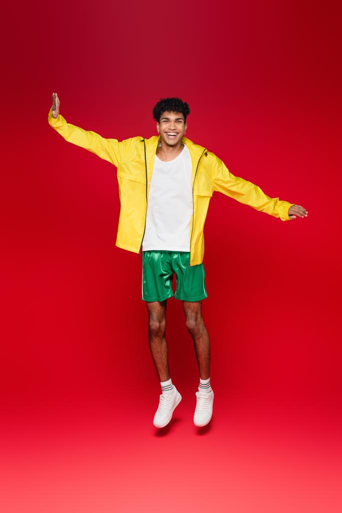 full length of happy african american man in yellow jacket jumping and talking on smartphone on red - Photo, Image