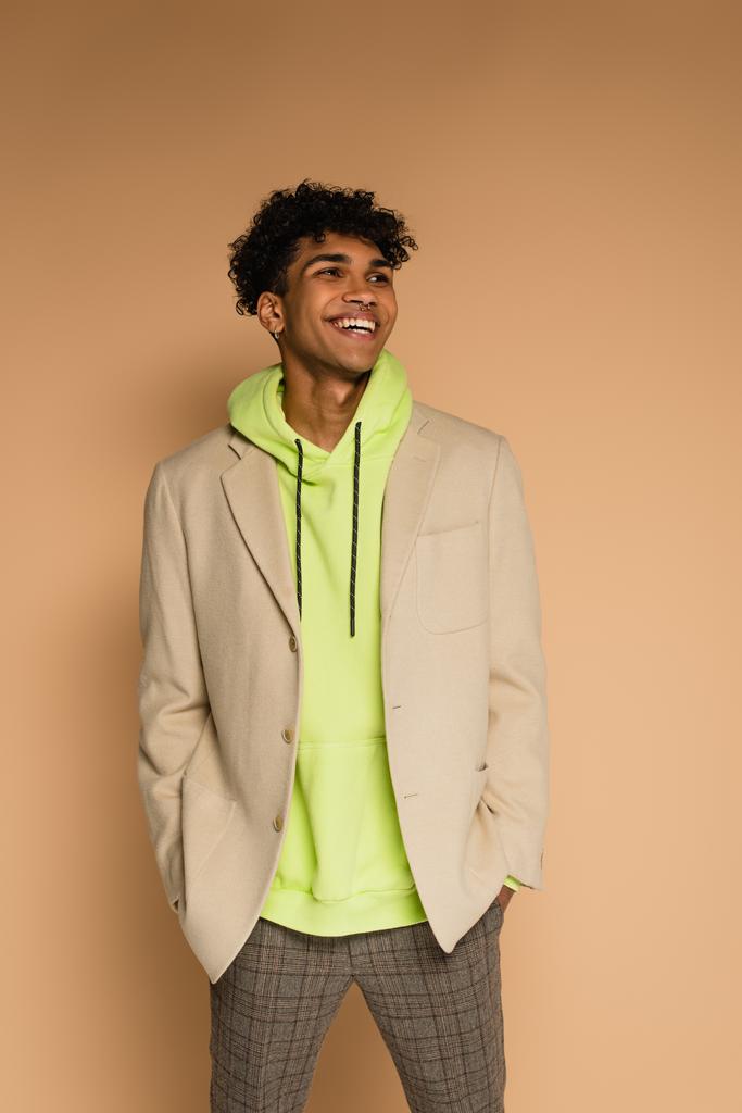 trendy uomo africano americano in felpa con cappuccio e blazer sorridente sul beige - Foto, immagini