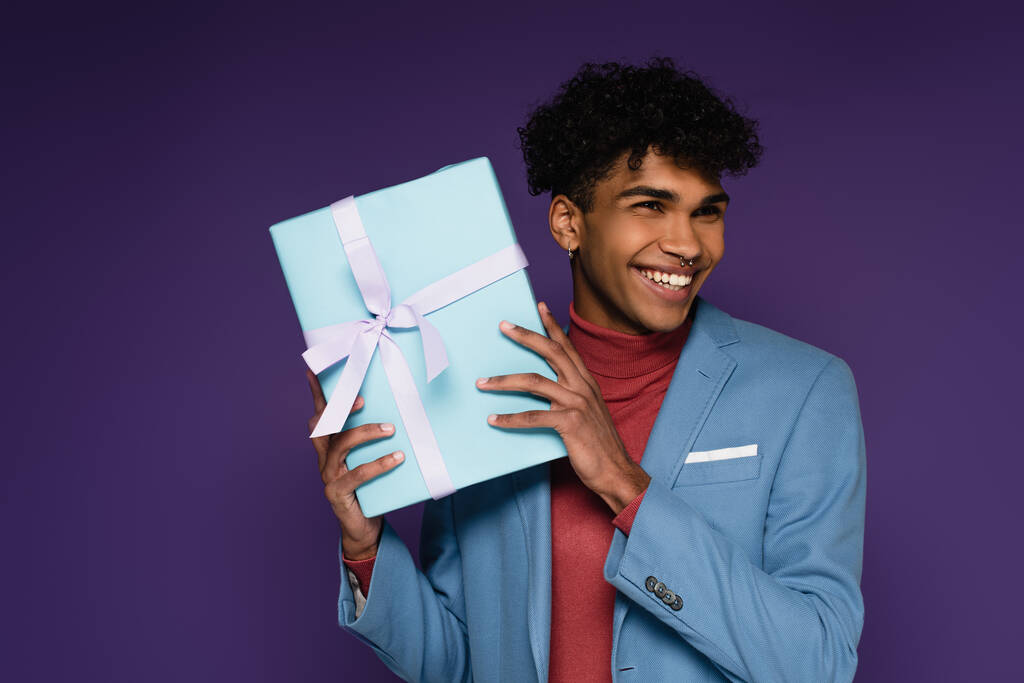 hombre afroamericano feliz en blazer azul sosteniendo regalo envuelto en púrpura - Foto, Imagen