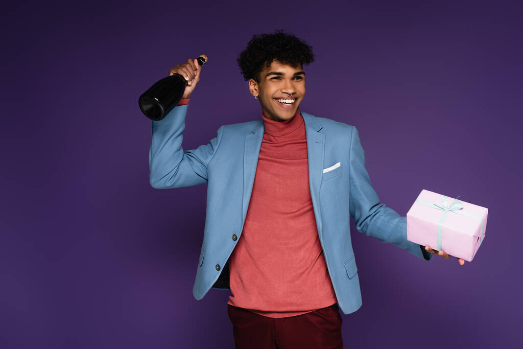 vrolijke Afro-Amerikaanse man in blauwe blazer met verpakt cadeau en fles champagne op paars - Foto, afbeelding