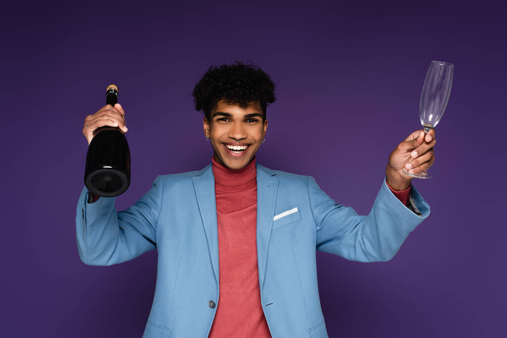 przekłuty Afrykański Amerykanin w niebieskiej marynarce uśmiecha się trzymając butelkę szampana na fioletowym - Zdjęcie, obraz