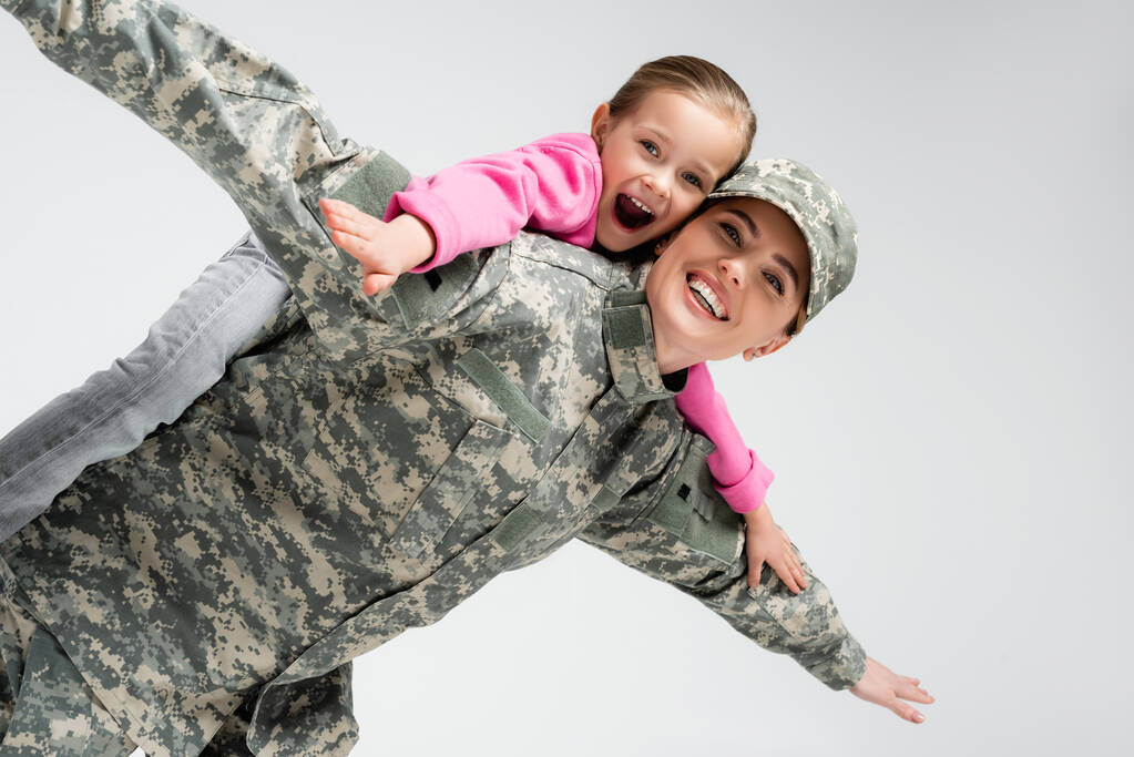Excited kid piggybacking on mother in military uniform isolated on grey  - Photo, Image
