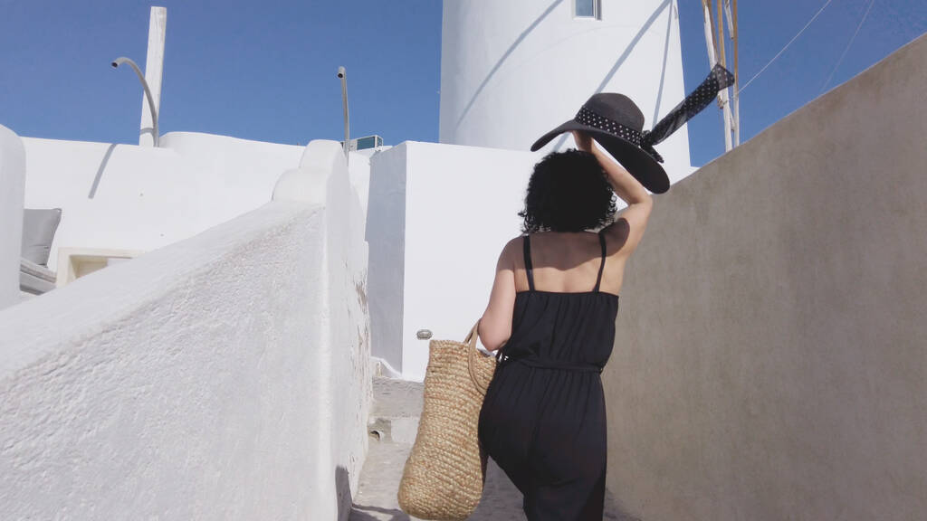 Mulher nova em uma roupa elegante preta e chapéu, andando na cidade de Oia, ilha de Santorini, Grécia, câmera lenta - Foto, Imagem