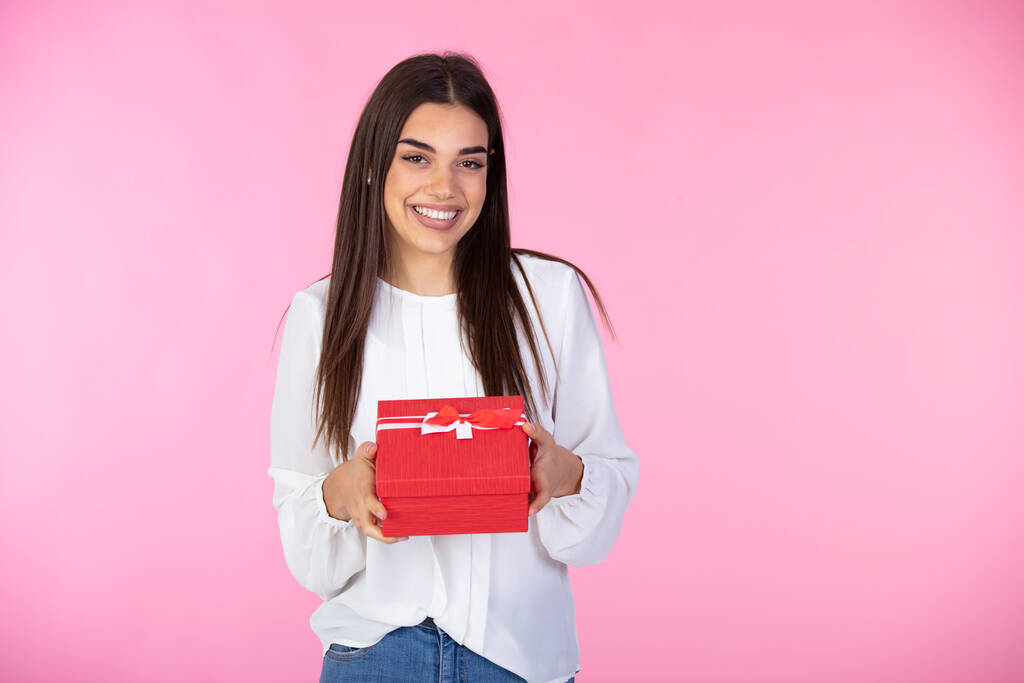 笑顔の女性は赤いギフトボックスを保持します。孤立したピンクの背景。スタジオだ。幸せな女性のイメージでシャツを保持ギフトボックスとピンクの背景の上にカメラを見て - 写真・画像
