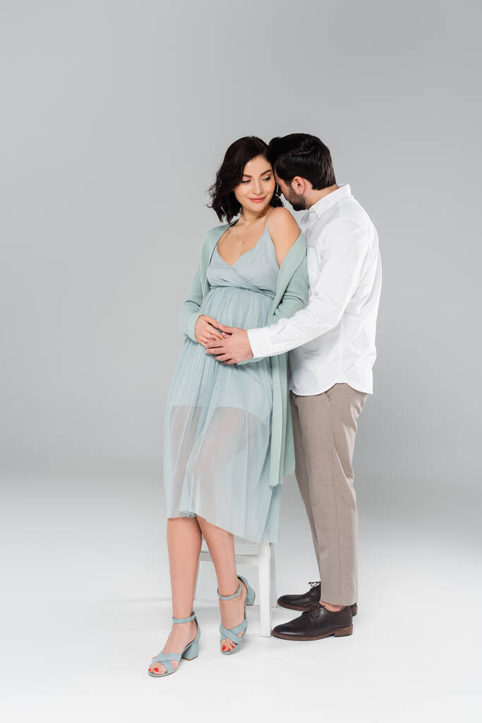 Мужчина обнимает беременную жену возле белого стула на сером фоне - Фото, изображение