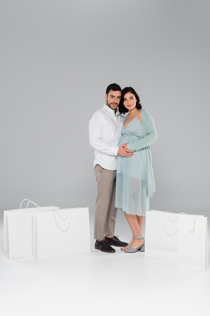 Smiling man hugging pregnant wife near white shopping bags on grey background - Photo, Image