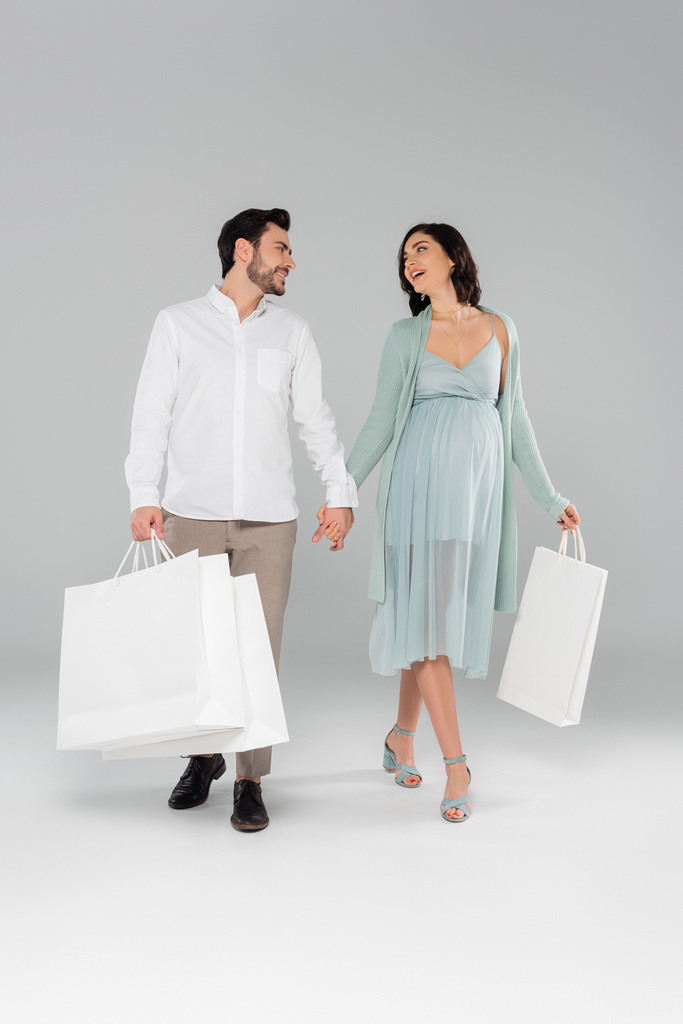 Mujer embarazada sosteniendo bolsa de compras y mano de marido mientras camina sobre fondo gris - Foto, Imagen