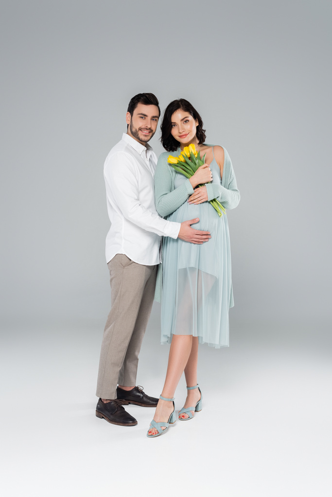 Fröhlicher Mann umarmt schwangere Frau im Kleid mit Blumen auf grauem Hintergrund - Foto, Bild
