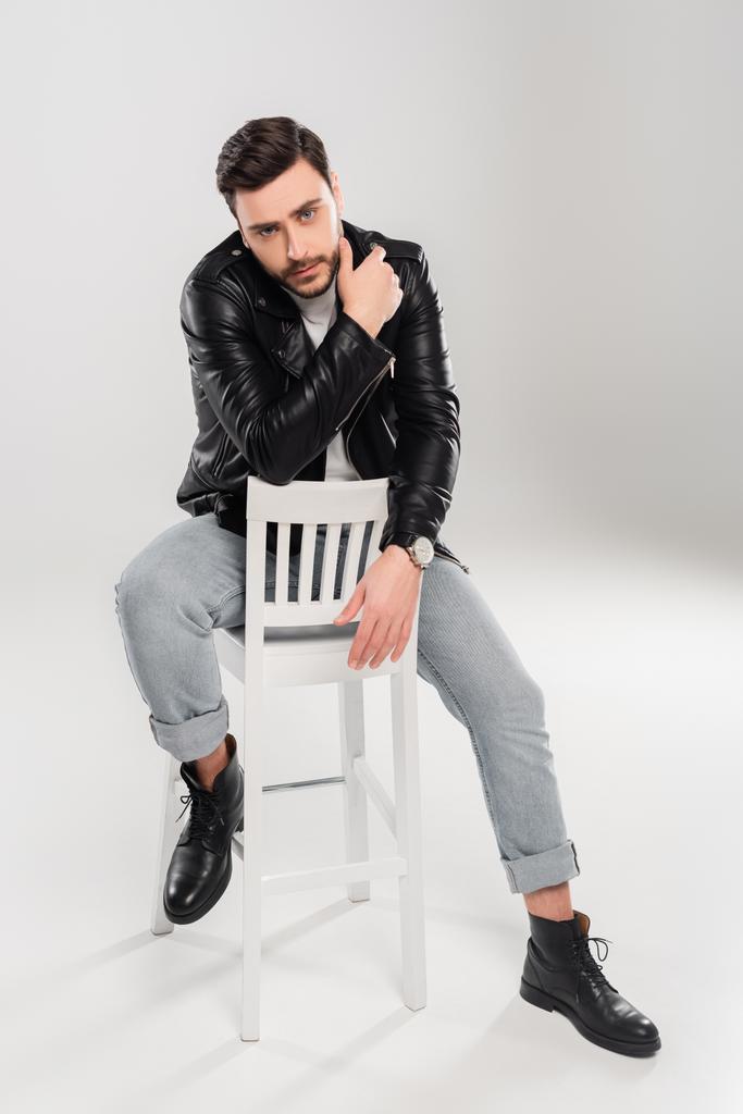 Бородатий стильний чоловік сидить на крихкому стільці на сірому фоні
  - Фото, зображення