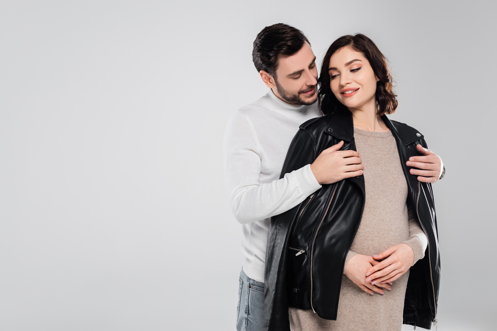 Homme souriant portant une veste sur jeune femme enceinte isolée sur gris  - Photo, image