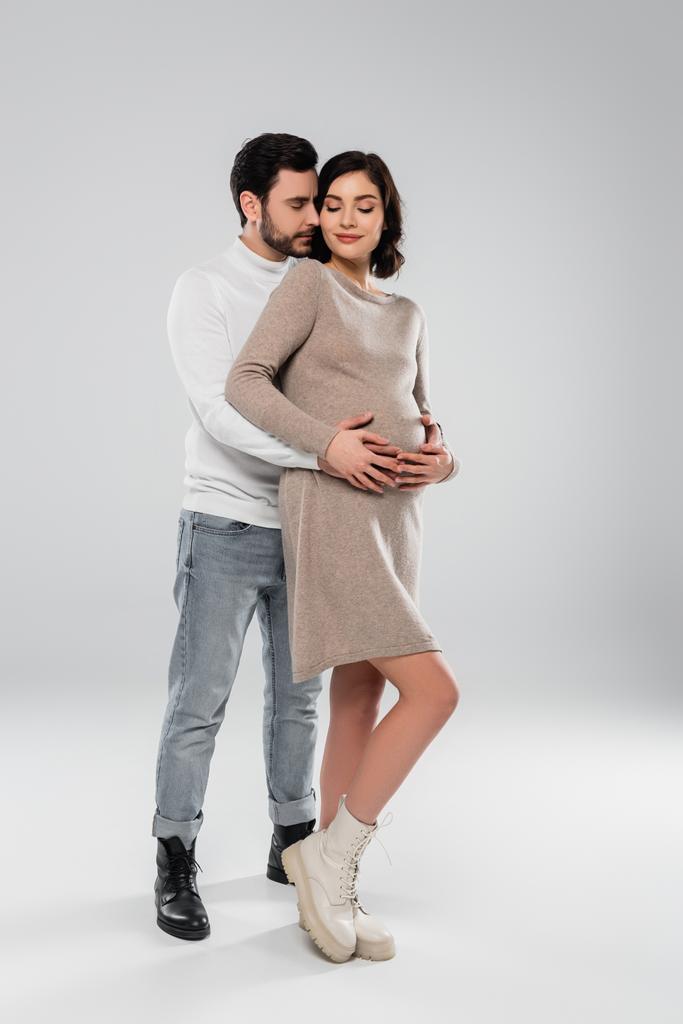 Stilvoller Mann umarmt lächelnde Schwangere in Kleid und Stiefeln auf grauem Hintergrund  - Foto, Bild