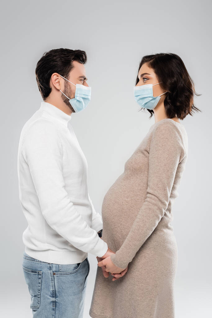 Вид збоку вагітної жінки в медичній масці, що тримає руки чоловіка ізольовані на сірому
  - Фото, зображення
