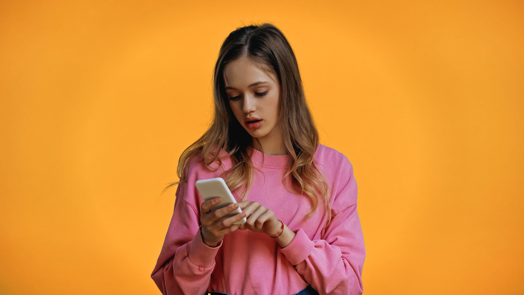 adolescente en sudadera rosa mensajería en el teléfono inteligente aislado en amarillo - Foto, Imagen