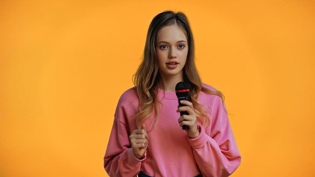 дівчина-підліток в рожевому светрі говорить, тримаючи мікрофон ізольовано на жовтому
 - Фото, зображення