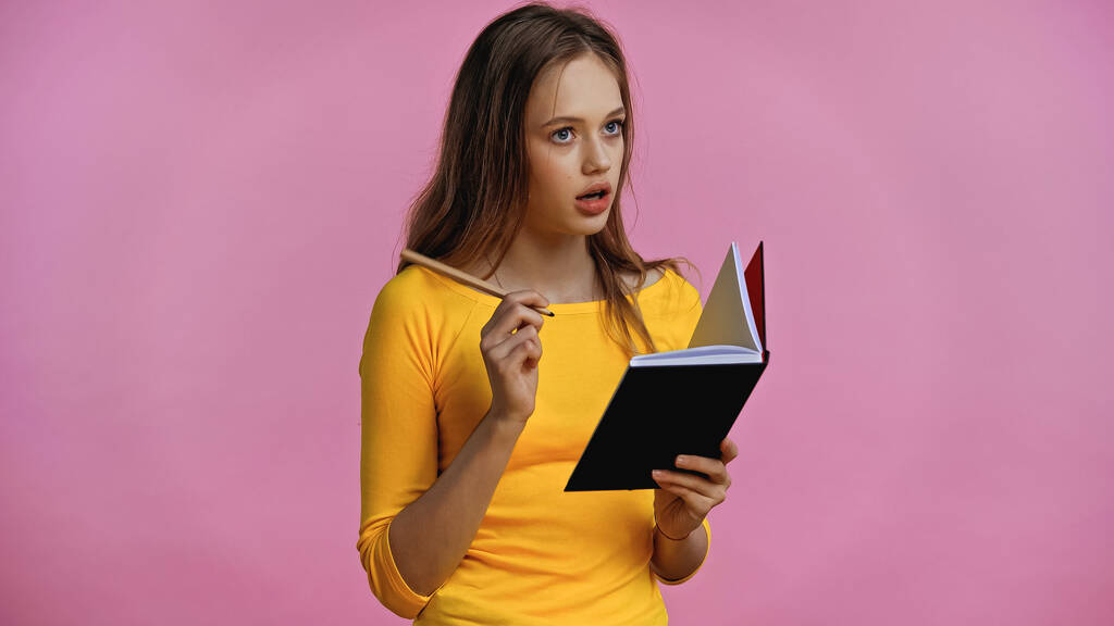 zamyšlená dospívající dívka drží notebook a pero izolované na růžové  - Fotografie, Obrázek