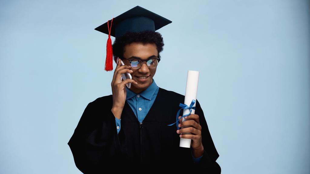 african american teenager in graduation gown and cap holding diploma while talking on smartphone isolated on blue - Photo, Image