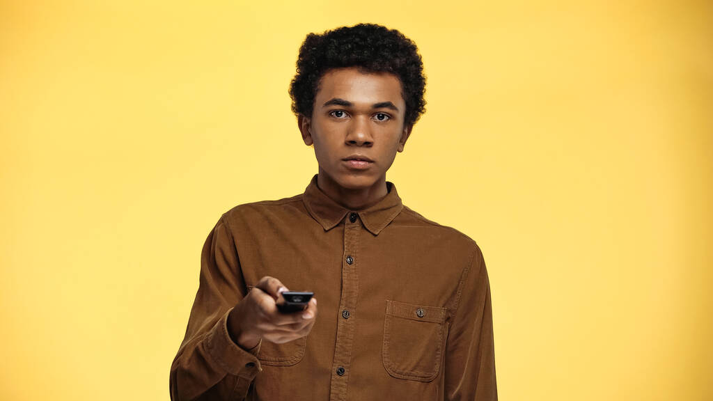 krullend afrikaans amerikaans tiener jongen houden afstandsbediening geïsoleerd op geel - Foto, afbeelding