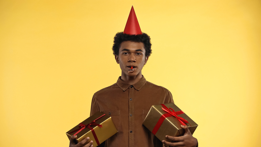 african american teenager in party cap holding gift boxes isolated on yellow - Photo, Image