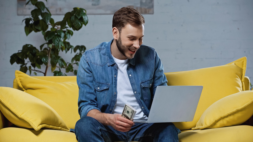 happy man holding dollars and using laptop while sitting on sofa - 写真・画像
