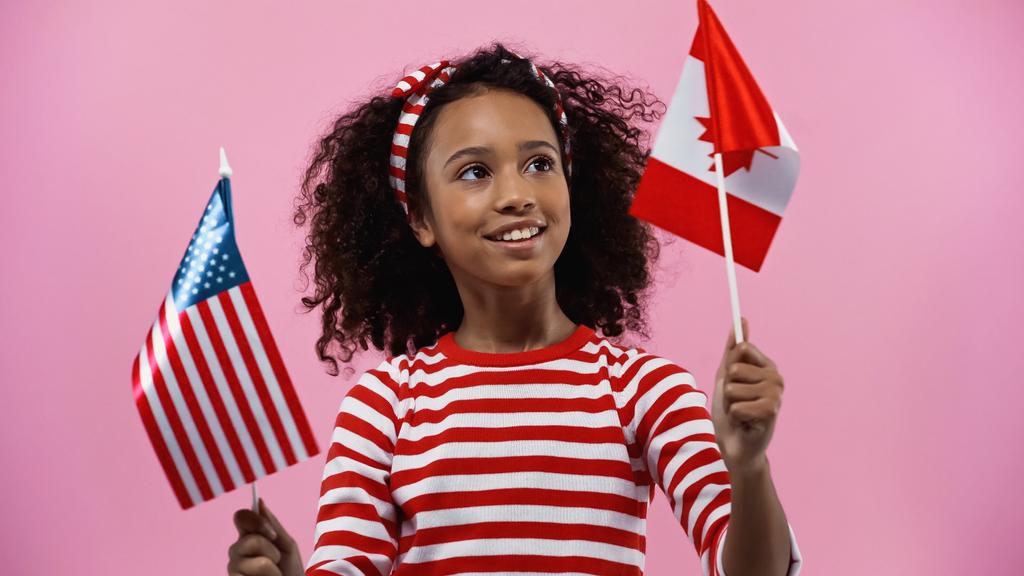жизнерадостная африканская американская девушка с флагами Америки и Канады, изолированными в розовом  - Фото, изображение