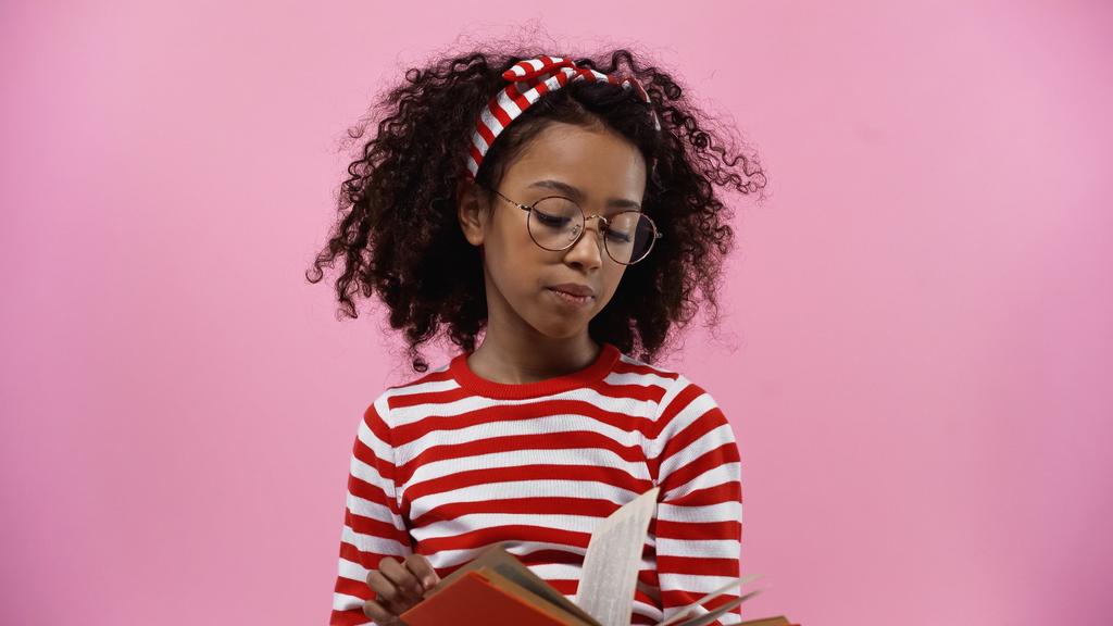 curly african american girl in glasses reading novel isolated on pink  - 写真・画像