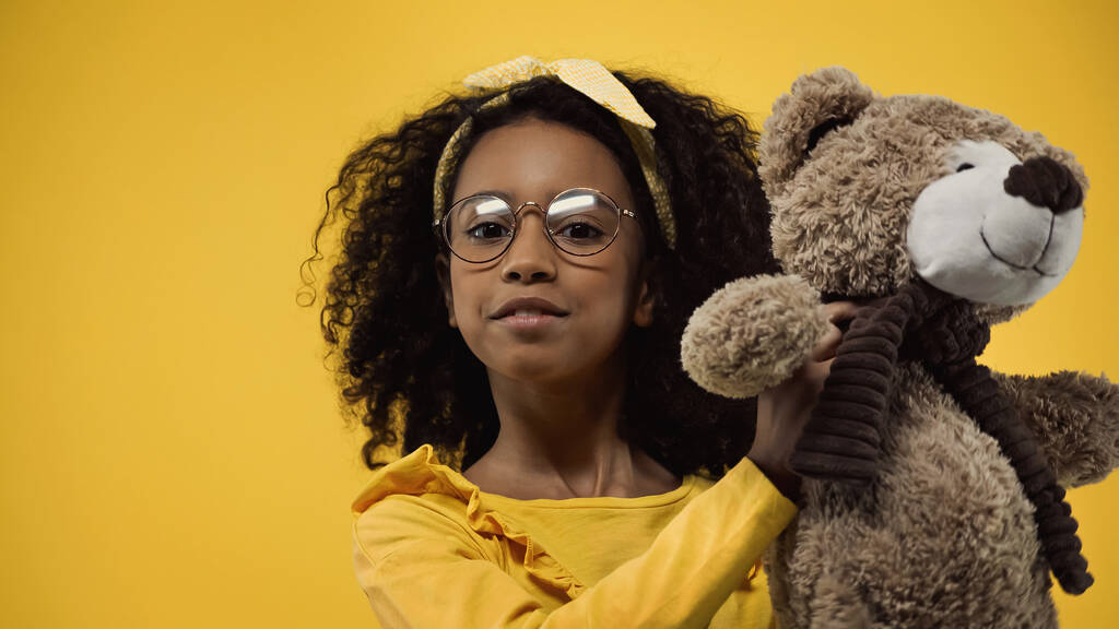 cacheados criança americana africana em óculos segurando ursinho isolado no amarelo - Foto, Imagem