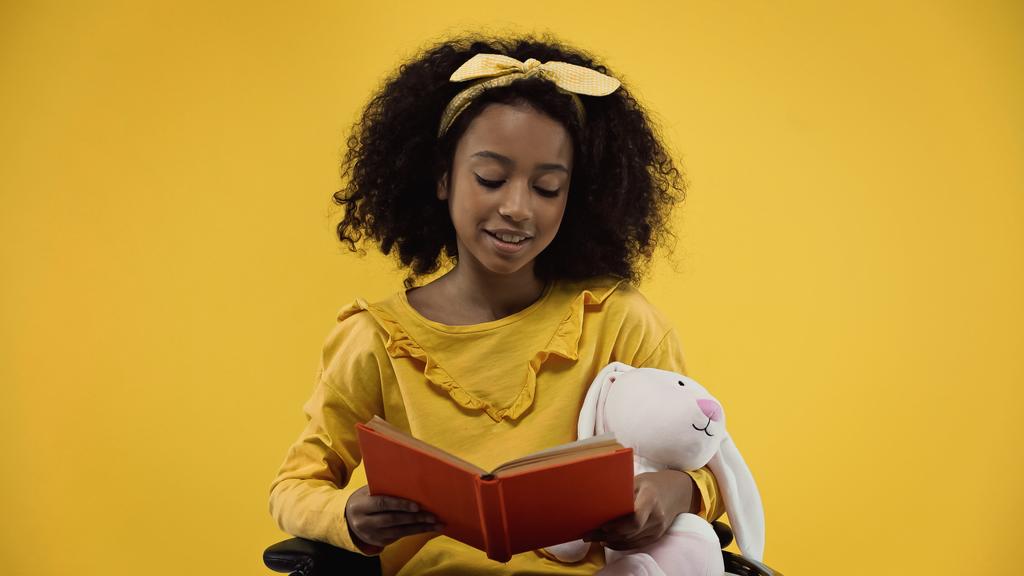 happy african american girl with soft toy reading book isolated on yellow - Photo, Image