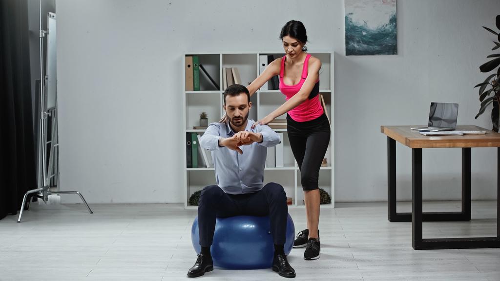 Entrenador de fitness de pie cerca de hombre de negocios en la pelota de fitness en la oficina  - Foto, Imagen