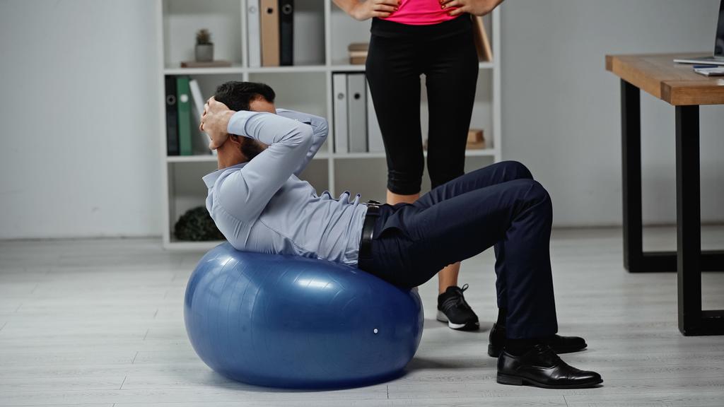 Fitnesstrainerin steht neben Geschäftsmann beim Training auf Fitnessball im Büro  - Foto, Bild