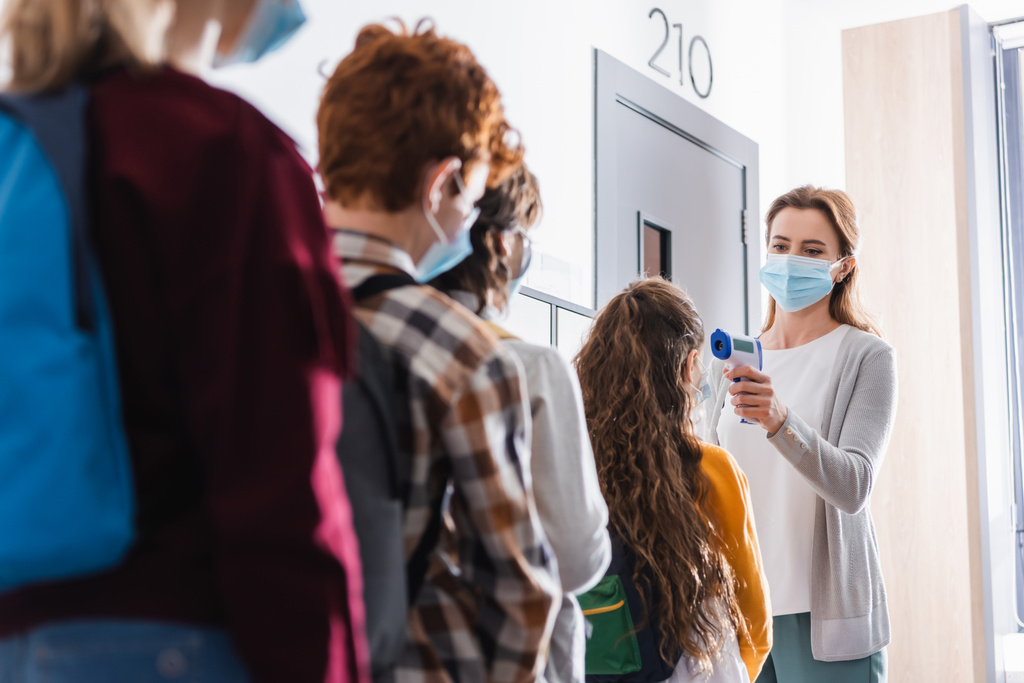 Professor em máscara protetora que mede a temperatura de estudante perto de alunos em primeiro plano borrado no corredor - Foto, Imagem