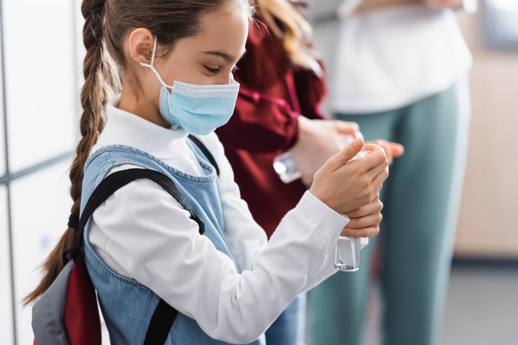 Studentessa in maschera medica utilizzando disinfettante mano vicino compagno di classe e insegnante su sfondo sfocato  - Foto, immagini