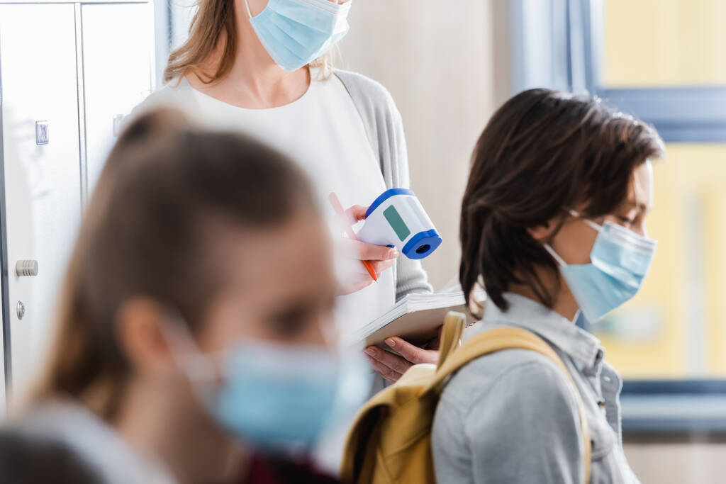 Učitel drží infračervený teploměr a notebook v blízkosti žáků v lékařských maskách na rozmazaném popředí  - Fotografie, Obrázek